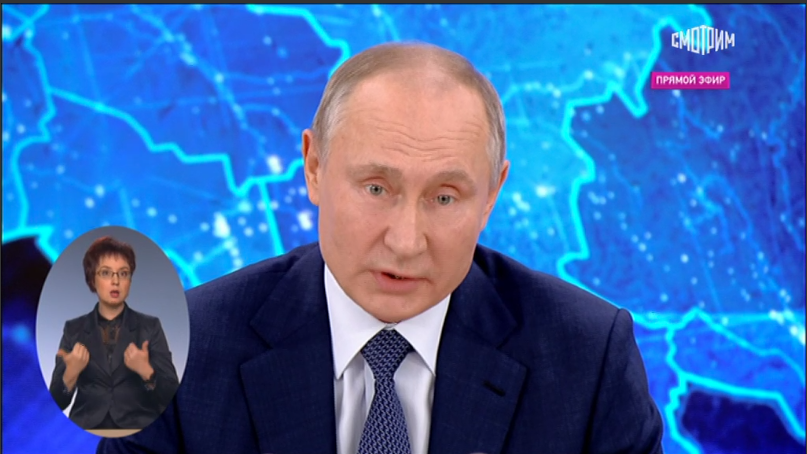 Картинка цветная. Фон похож на карту, но только карта в синем и голубом цвете. По середине картинки сидит Президент Российской Федерации Владимир Владимирович Путин. На нем белая рубашка, галстук синий в горошек и синий пиджак. Слева от Владимира Владимировича, в отдельном окне в форме овала, находиться женщина. Она – сурдопереводчик. У неё короткая стрижка, каштановые волосы. На ней синий пиджак.