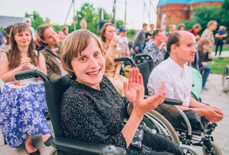 Цветная фотография. На фотографии много людей, они находятся на улице летом. На первом плане девушка хлопает в ладоши. У неё с короткой стрижкой и светлыми волосами. На ней черная кофта. Сзади неё также есть люди.