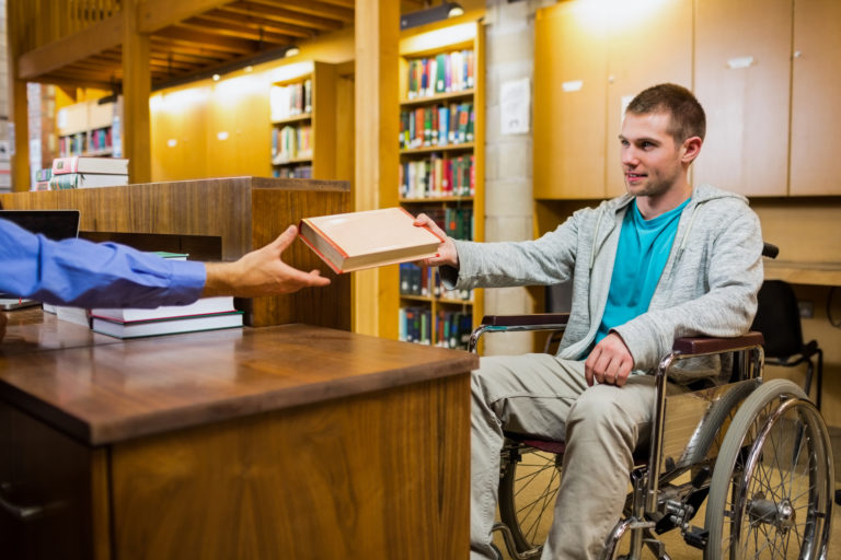 Фото цветное. Это библиотека. Много вещей из дерева. Много полок, шкафов. На переднем плане мужчина на коляске. У него короткая стрижка, русые волосы. На нем голубая футболка, сверху серая кофта и брюки. Он протягивает книгу правой рукой мужчине в синей рубашке. Перед ним стоит стол.