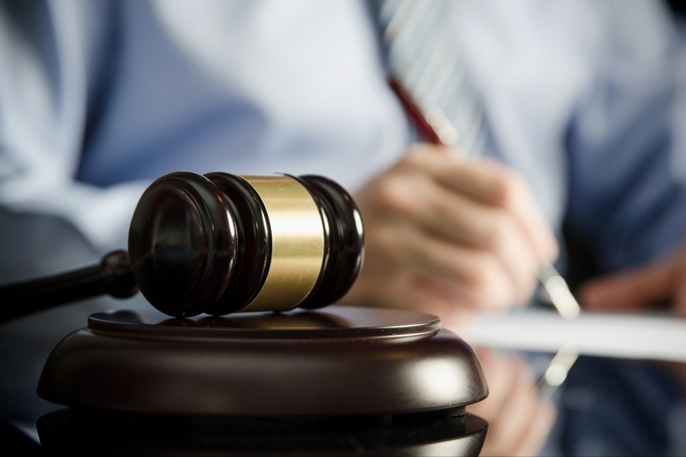 Фото цветное. Мужчина в голубой рубашке с галстуком в полосочку. Он заполняет бумаги коричневой ручкой. Его изображение размыто. Перед ним судейский молоток.