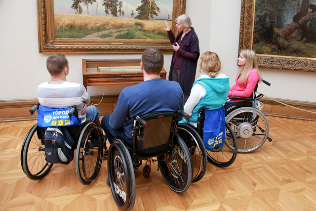 Фото цветное. Комната светлая. Четыре человека на коляске находятся в музее. Перед ними стоит экскурсовод. Она рассказывает и показывает о картине. На картине нарисовано поле из пшенице, посередине дорожка, стоят одиночные деревья. 