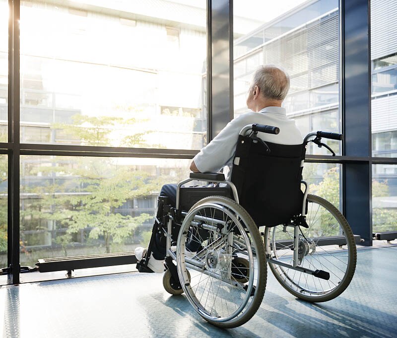 Фото цветное. В коляске сидит мужчина. У него седые волосы. На нем белая футболка и черные брюки. Он смотрит в окно. Окна очень большие, в них видно деревья и другую часть больницы.