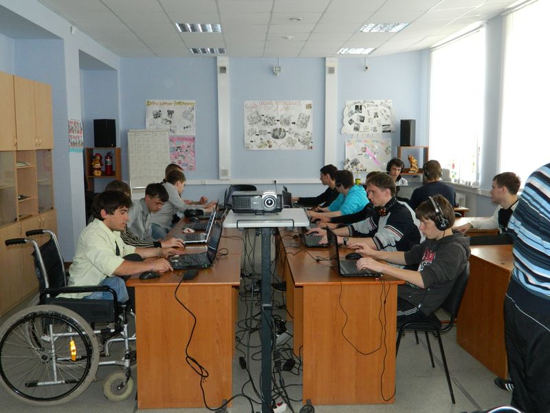 Цветное фото. Комната, напоминающая учебный класс. В середине комнаты находятся столы, придвинутые друг к другу. За ними сидят студенты за компьютерами, по центру находятся проектор. Вдоль стен шкафы, на одной из стен прикреплены плакаты, напоминающие коллажи.