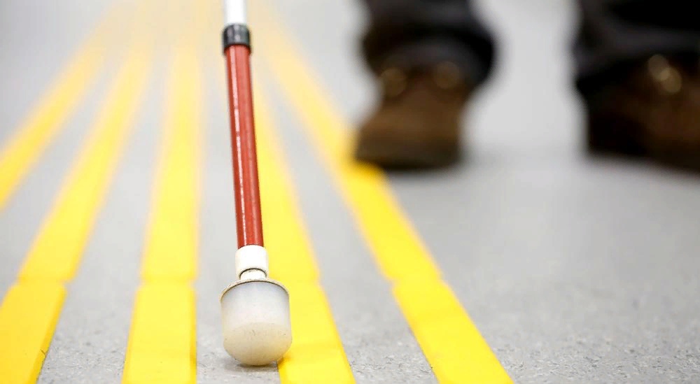 Фото цветное. Слева пять жёлтых полосок. По ней ведут трость. Она красного цвета. Её ведет мужчина. На нем темные брюки и коричневые ботинки. На фото видно только его ноги. 